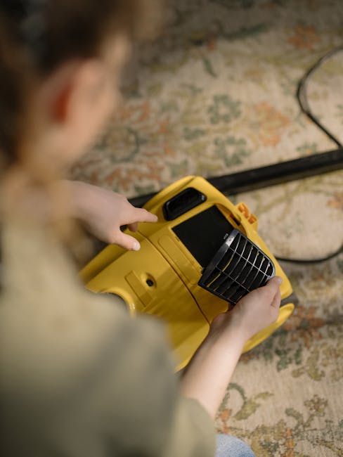 A person using a yellow and black portable vacuum cleaner on a patterned carpet, with hands adjusting the cleaner's dust compartment, in a room illuminated by natural light. The vacuum cleaner's hose