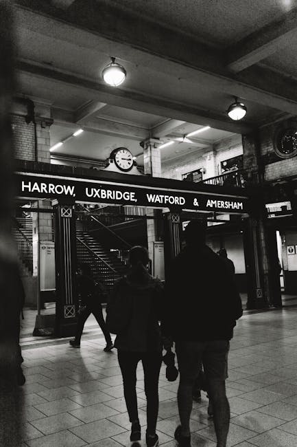 A black-and-white photograph of the Harrow Uxbridge, Watford, and Amersham underground station entrance, featuring a large illuminated sign with station names, a round ceiling light, and a clock mounted on the ceiling. In the foreground, two silhouetted travelers stand together, with other passengers visible walking in the station. The station's modern design includes tiled floors, metal staircases, and bright lighting, creating a clean and organized environment. The overall scene highlights the station's well-maintained and hygienic appearance, emphasizing the importance of surface cleaning and maintenance for a safe public space, as promoted by West Harrow Carpet Cleaners' professional cleaning services near Harrow on the Hill station.