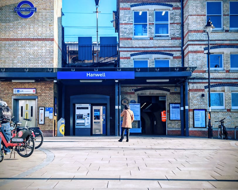 A woman dressed in a beige coat and black trousers stands on a paved sidewalk outside Hanwell station in West Harrow, London, looking at ticket machines and information boards near the entrance. The station building features a brick facade with large blue signage displaying 'Hanwell' and the Elizabeth Line logo, and has multiple windows with white frames. There are bicycles parked to the right, a streetlamp, and a notice board with station-related information mounted on the brick wall. The scene is illuminated by natural daylight, with a clear sky and some reflections visible on the ticket machines, presenting a clean and well-maintained urban setting.
