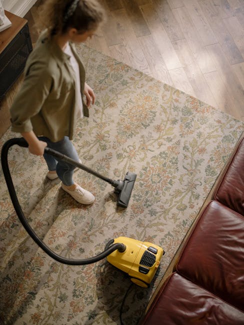 A person wearing beige shoes and grey jeans operates a carpet cleaning vacuum cleaner on a decorative, floral patterned area rug in a domestic setting. The vacuum cleaner is yellow with a black hose,
