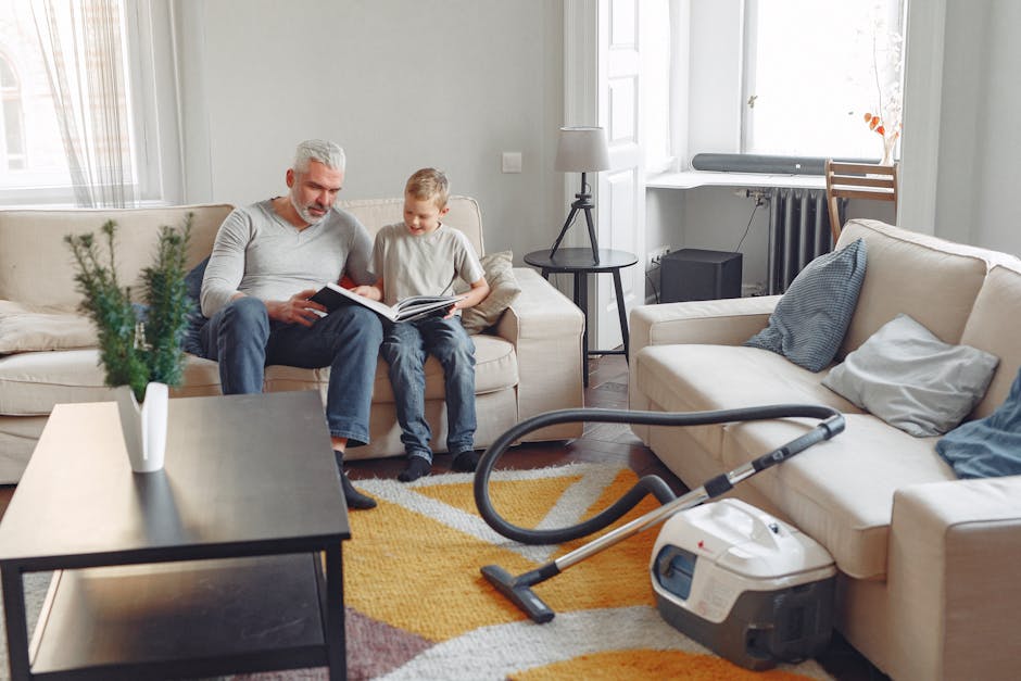 A living room with light-colored sofas and a patterned area rug, featuring a vacuum cleaner parked on the rug near a cream sofa. An older man and a young boy sit on another sofa, reading a book togeth