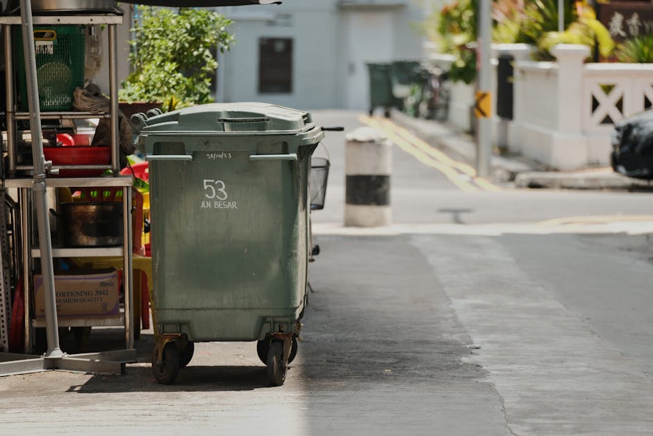 A street scene featuring a sanitation worker dressed in a bright green uniform and cap, collecting waste from a large green wheeled bin placed on a paved sidewalk. The worker is leaning into the bin, 